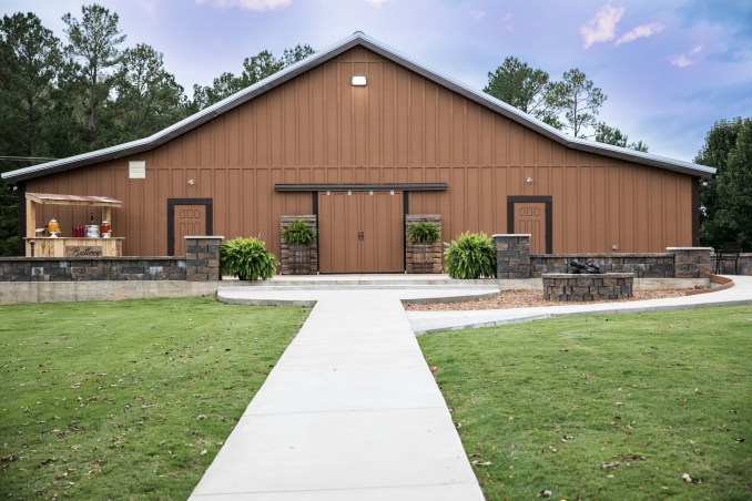 Brown barn-style building featuring entrance doors framed by potted plants, with a paved path leading up to it, set in a green landscape.