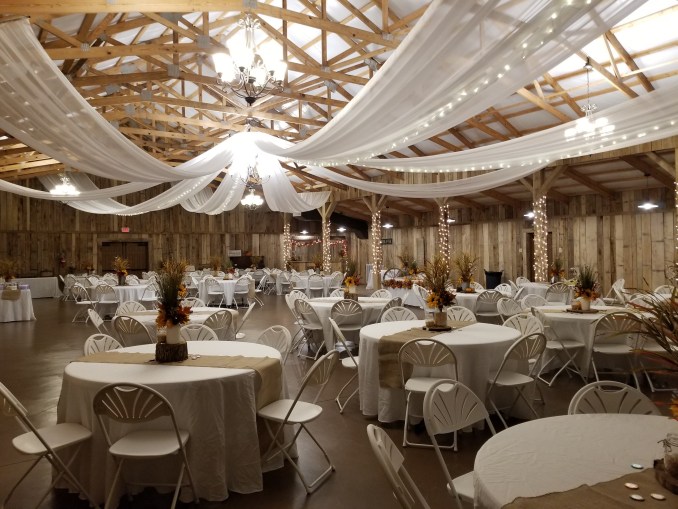 Elegant event space featuring round tables with white tablecloths and floral centerpieces under draped fabric and chandeliers, perfect for celebrations.