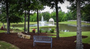 Serene pond scene featuring a fountain and gazebo, surrounded by trees and landscaped beds, with a blue bench offering a relaxing view.