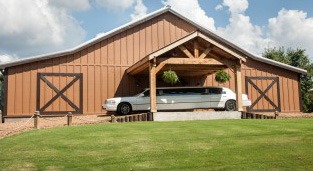 A white limousine parked under a wooden overhang at a spacious barn, set against a sunny blue sky, highlighting a unique event venue.