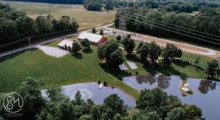 Aerial view of a scenic rural property featuring a horse riding area, a red barn, grassy fields, and a large pond, surrounded by trees.