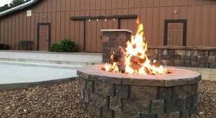 A stone fire pit with a vibrant flame, situated in front of a brown building, creating a cozy outdoor gathering space.