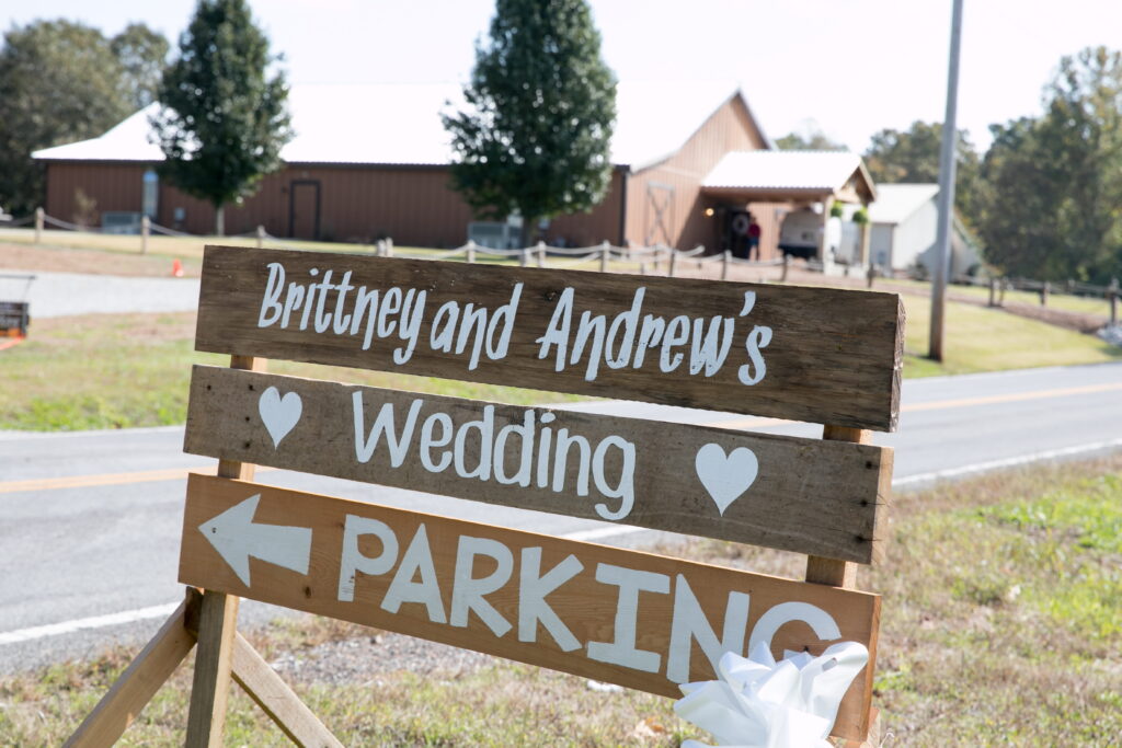 Wooden sign directing guests to Brittney and Andrew's wedding parking, featuring decorative hearts and arrows, with a rustic venue in the background.