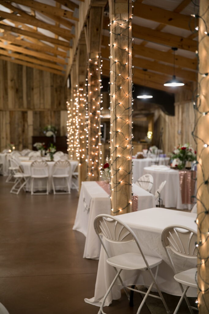 Rustic event space with wooden beams and twinkling fairy lights. Tables are set with white linens, creating an inviting atmosphere for gatherings.