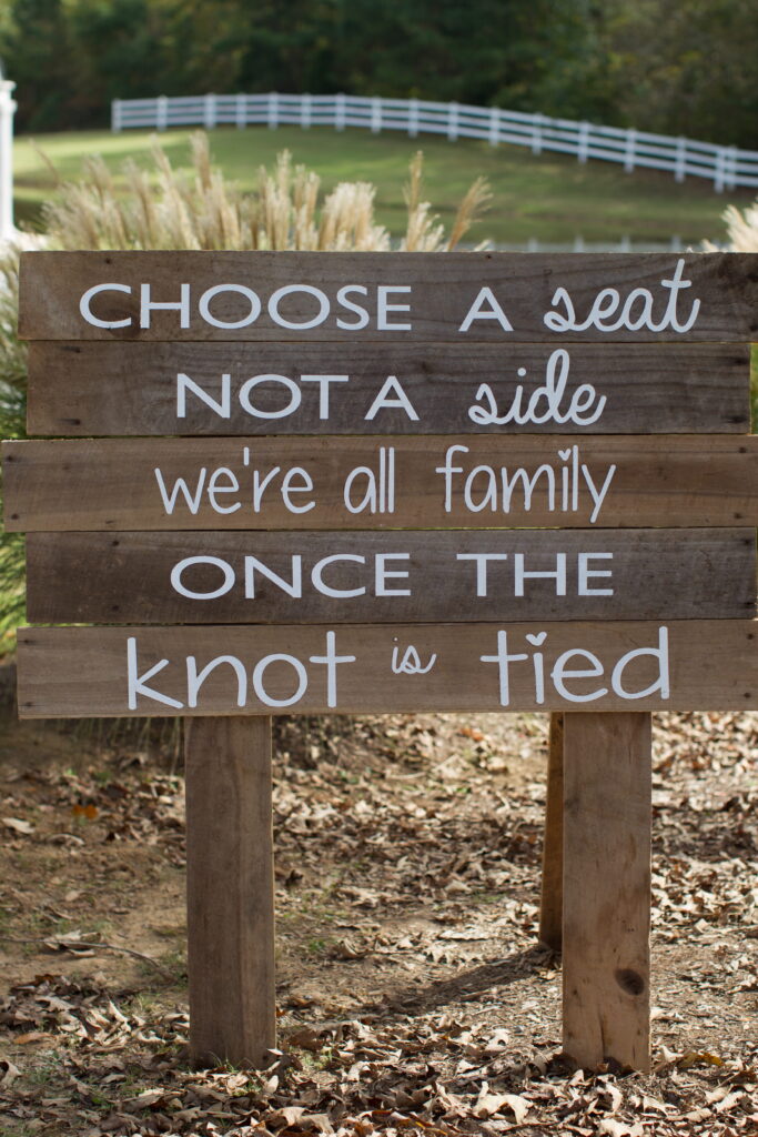 Wooden sign reads, "Choose a seat, not a side. We're all family once the knot is tied." It conveys a welcoming message for wedding guests.