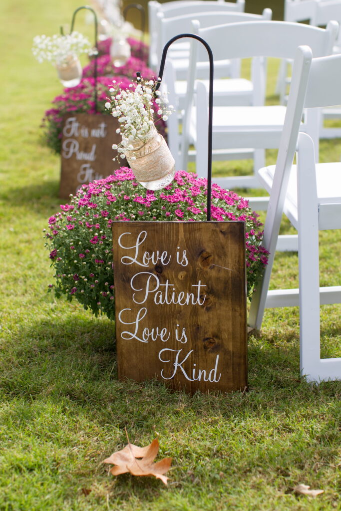 Wooden sign displays the phrases "Love is Patient" and "Love is Kind," surrounded by pink flowers and wedding seating, enhancing a romantic outdoor setting.