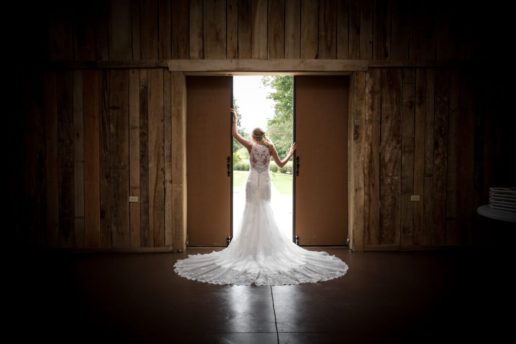 Bride in a wedding gown stands poised at the open barn doors, her trailing veil cascading behind her, framing a serene outdoor scene.