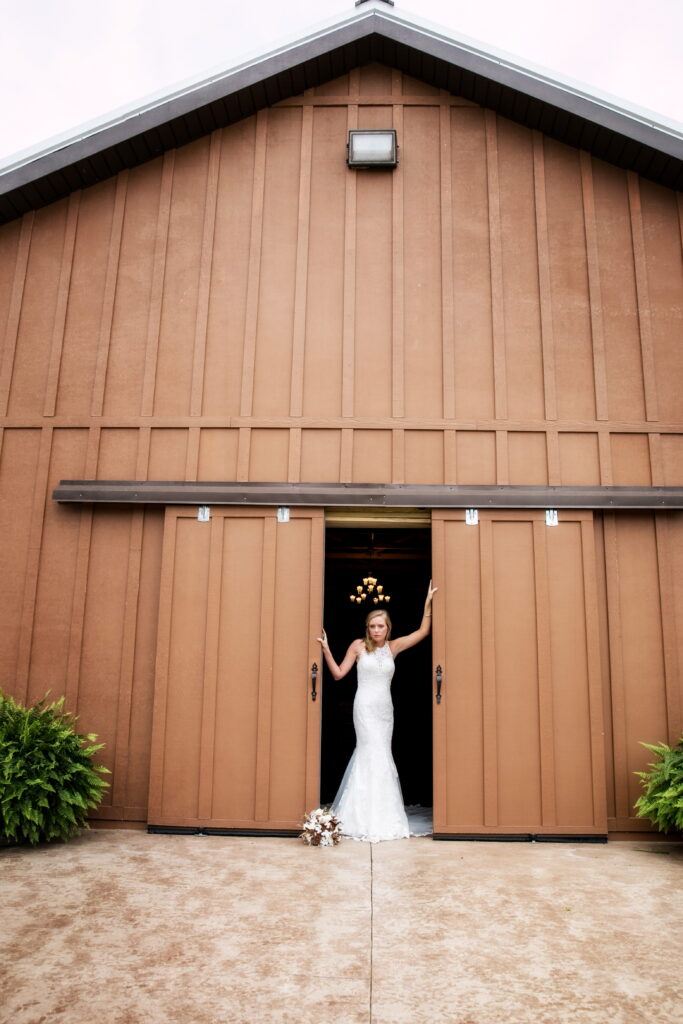 Bride in a stunning lace gown stands at the entrance of a brown barn, holding a bouquet and posing gracefully. Perfect for wedding-related content.