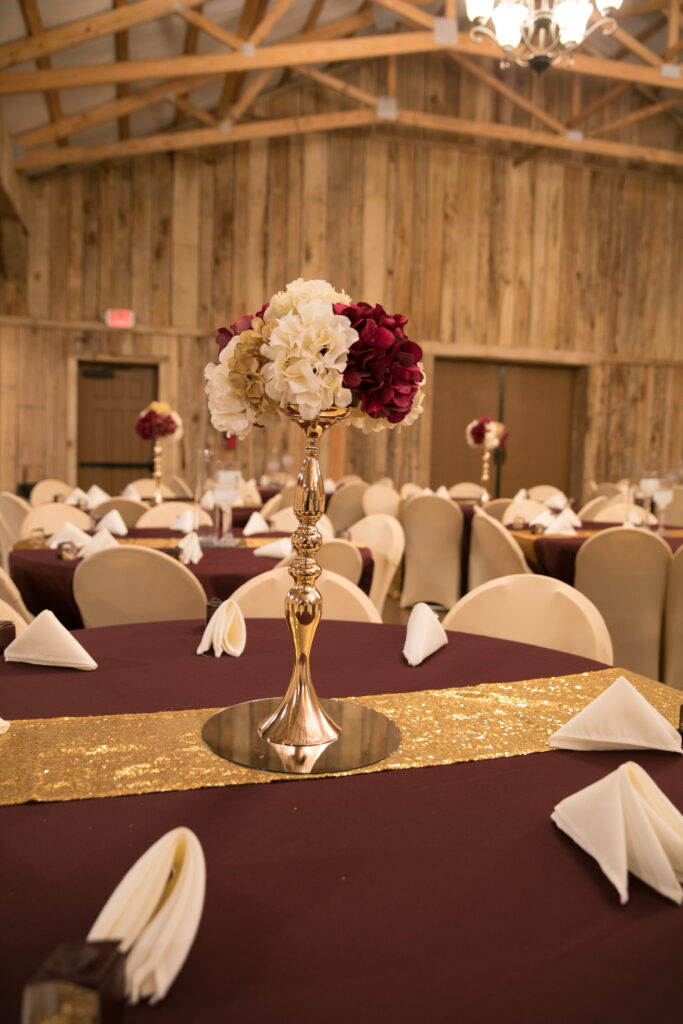 Elegant table setting in a rustic venue, featuring a tall floral centerpiece with burgundy and white blooms, accented by golden table runners.