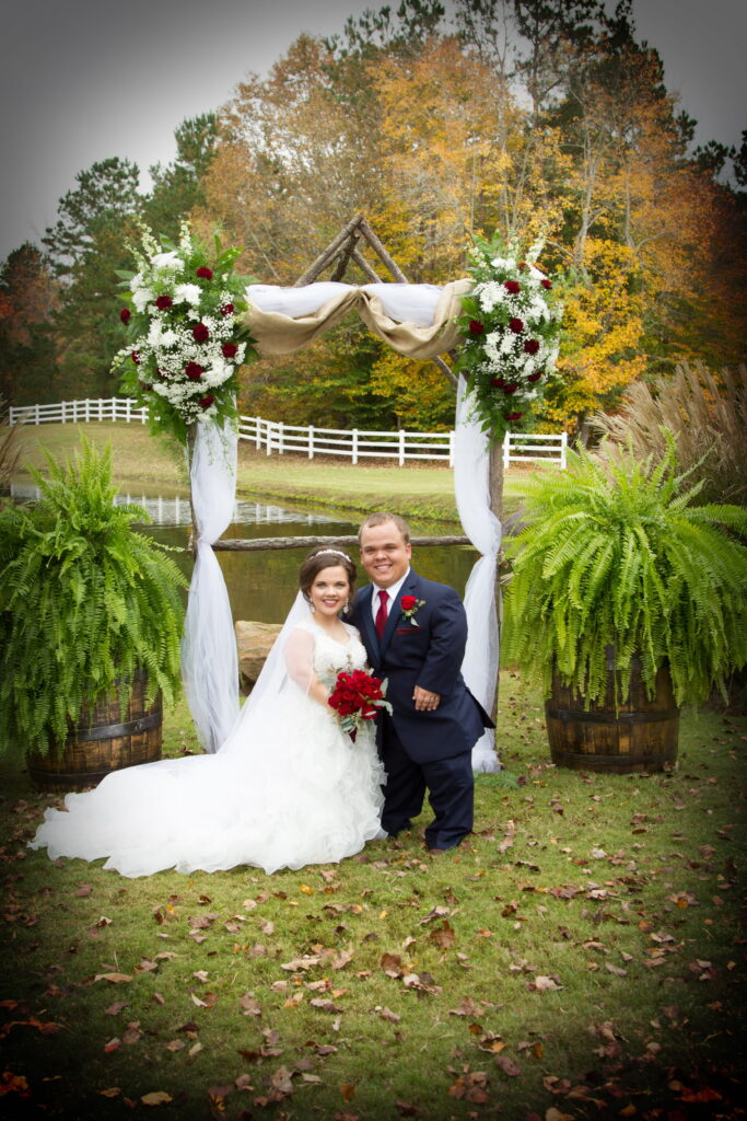 Bride and groom pose together under a floral arch at their outdoor wedding, surrounded by autumn foliage and a serene pond.