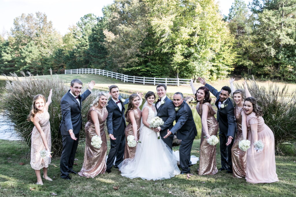 A joyful wedding party poses by a pond, showcasing the bride and groom at the center, surrounded by friends in elegant attire against a natural backdrop.