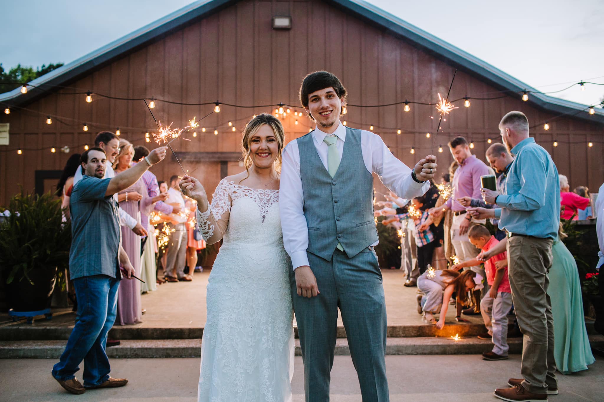 A couple kisses surrounded by wedding guests holding sparklers, celebrating their special moment outside a venue at dusk.