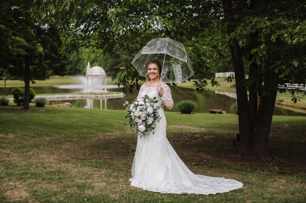 Bride in a lace gown holds a clear umbrella and bouquet of white roses, standing in a lush garden by a pond with a fountain.