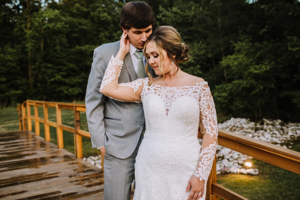 A couple embraces on a wooden bridge surrounded by greenery. The bride wears an elegant white lace gown, while the groom dons a light gray suit.