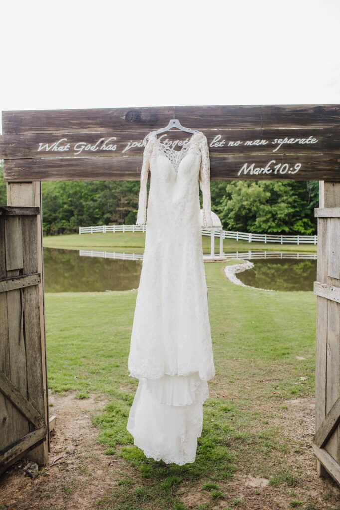 A wedding dress hangs on a wooden sign near a tranquil pond, featuring a quote from Mark 10:9 about marriage unity.