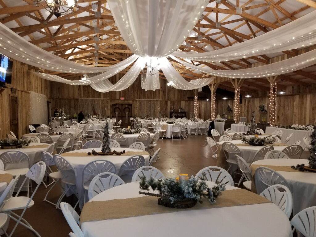 Rustic event space with wooden beams and draped fabric, featuring round tables set with holiday decor, creating a cozy atmosphere for gatherings.