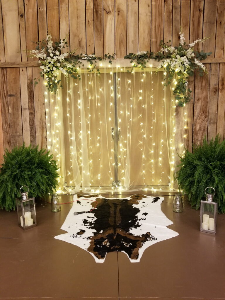 Rustic wedding backdrop featuring twinkling fairy lights, floral arrangements, and a cowhide rug, creating a warm and inviting atmosphere.