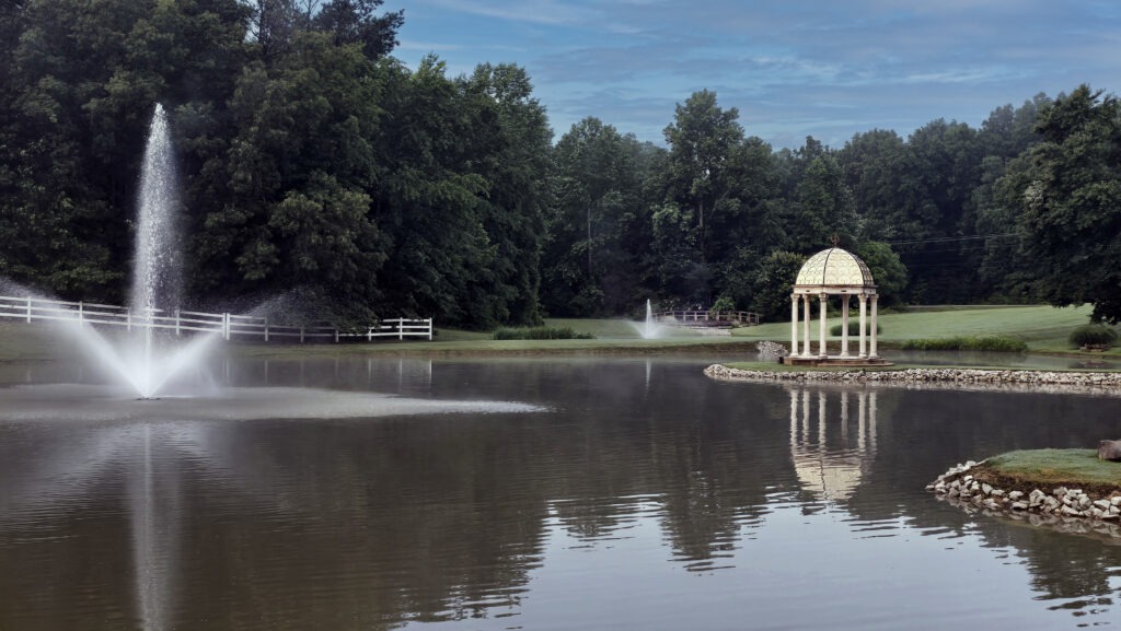 A serene pond features a decorative gazebo and a fountain, surrounded by greenery. The image captures a tranquil outdoor setting, ideal for relaxation.