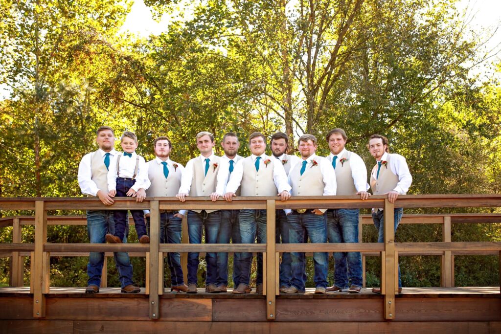 Groom and groomsmen pose on a wooden bridge, dressed in white shirts and beige vests, with a young boy in the front. Greenery surrounds them.