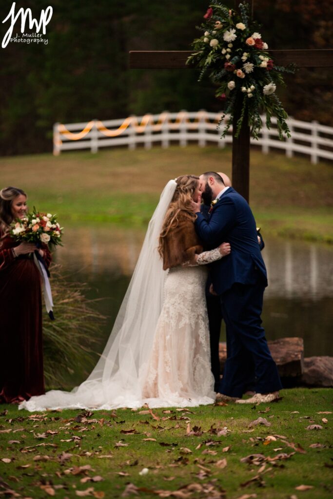 Bride in a lace gown and groom in a tuxedo share a romantic kiss outdoors, framed by autumn foliage and a serene pond in the background.