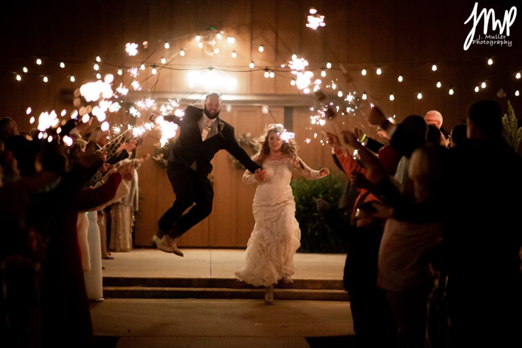 A joyful bride and groom exit their wedding, surrounded by waving guests holding sparklers, creating a festive atmosphere under twinkling lights.