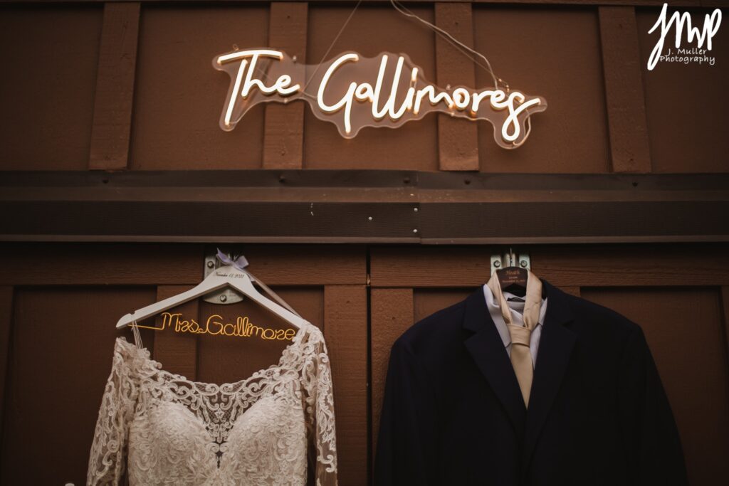 Wedding attire hangs against a wooden backdrop, featuring a lace wedding dress and a groom's suit, with a neon sign reading "The Gallimores."