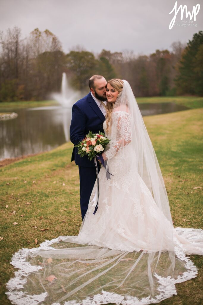 Bride and groom embrace by a tranquil pond, with a fountain in the background. The bride wears an elegant lace gown and holds a bouquet.
