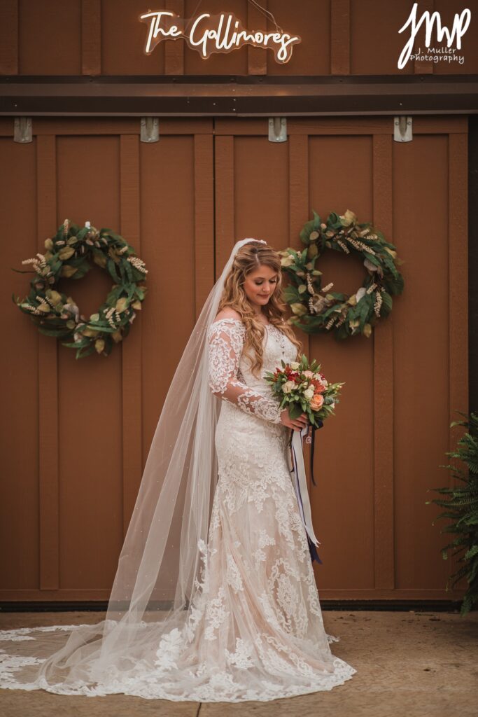 Bride in a lace wedding gown holds a bouquet in front of a rustic backdrop adorned with wreaths, celebrating her marriage to "The Gallimores."