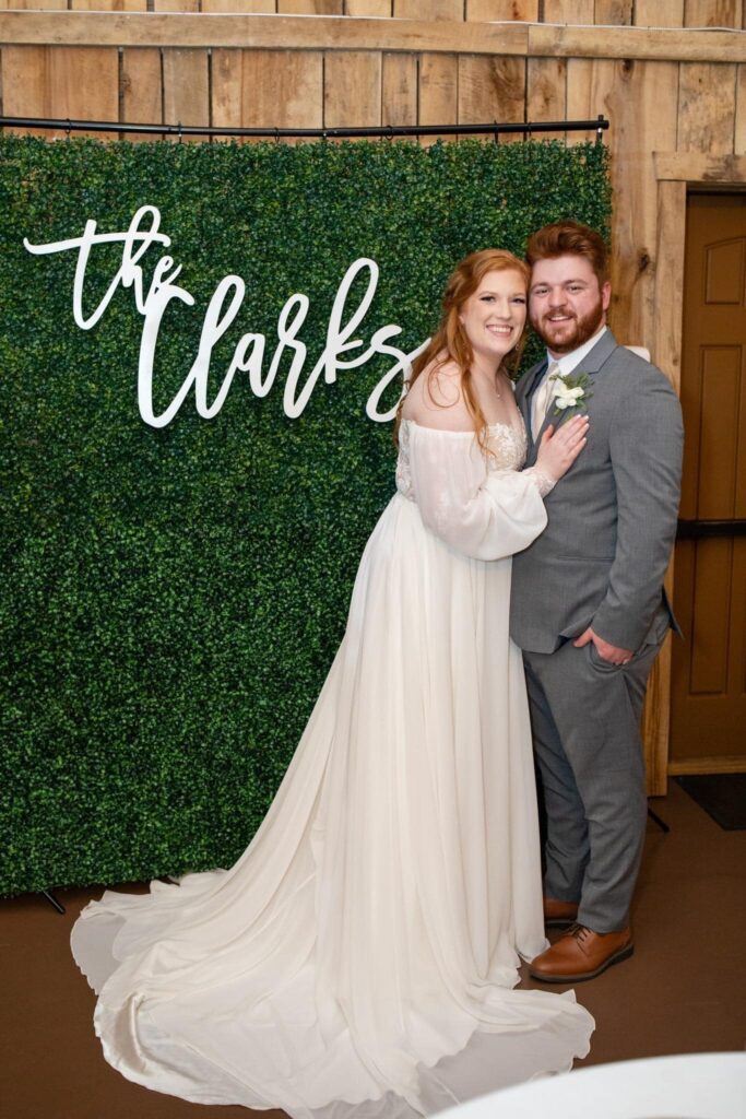 Bride and groom pose happily in front of a green floral backdrop with "the Clarks" sign. Celebrating their wedding day.
