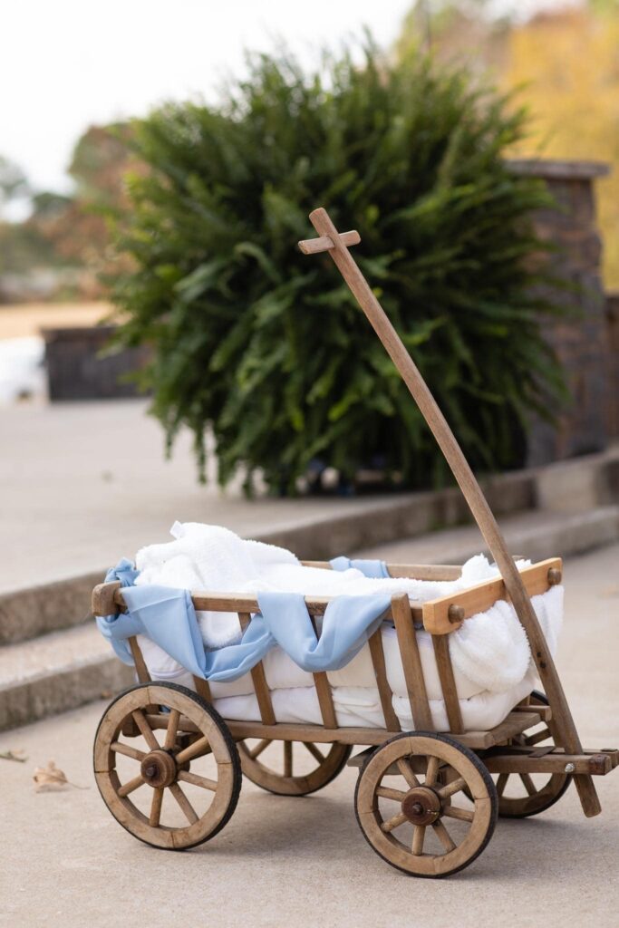 A rustic wooden cart draped with soft white blankets and blue fabric, standing against a backdrop of lush greenery, evokes warmth and charm.