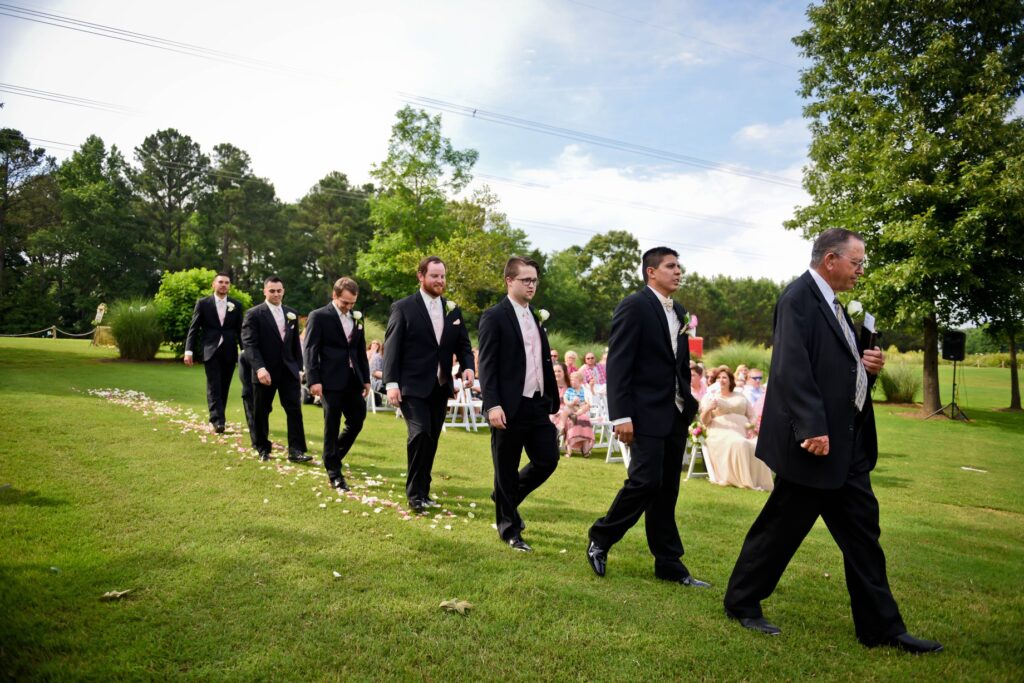 Groomsmen and the officiant walk down a grassy aisle, surrounded by guests, in a sunny outdoor wedding ceremony.