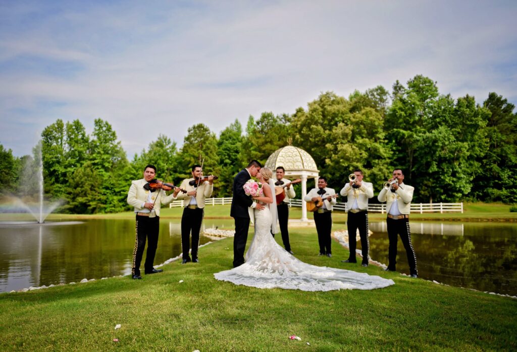 Bride and groom embrace by a pond, surrounded by a mariachi band playing instruments. A decorative gazebo and lush greenery create a romantic setting.