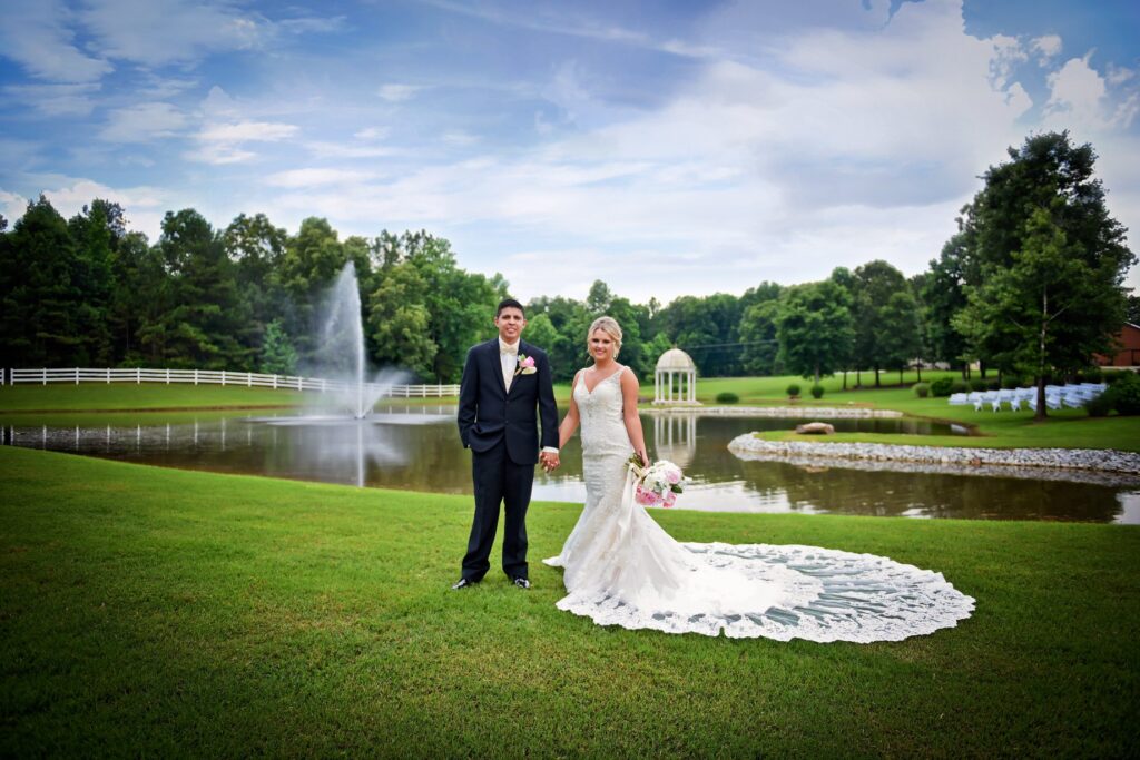 Bride and groom stand hand-in-hand by a serene pond with a fountain, surrounded by lush greenery, capturing their wedding moment in a scenic outdoor setting.