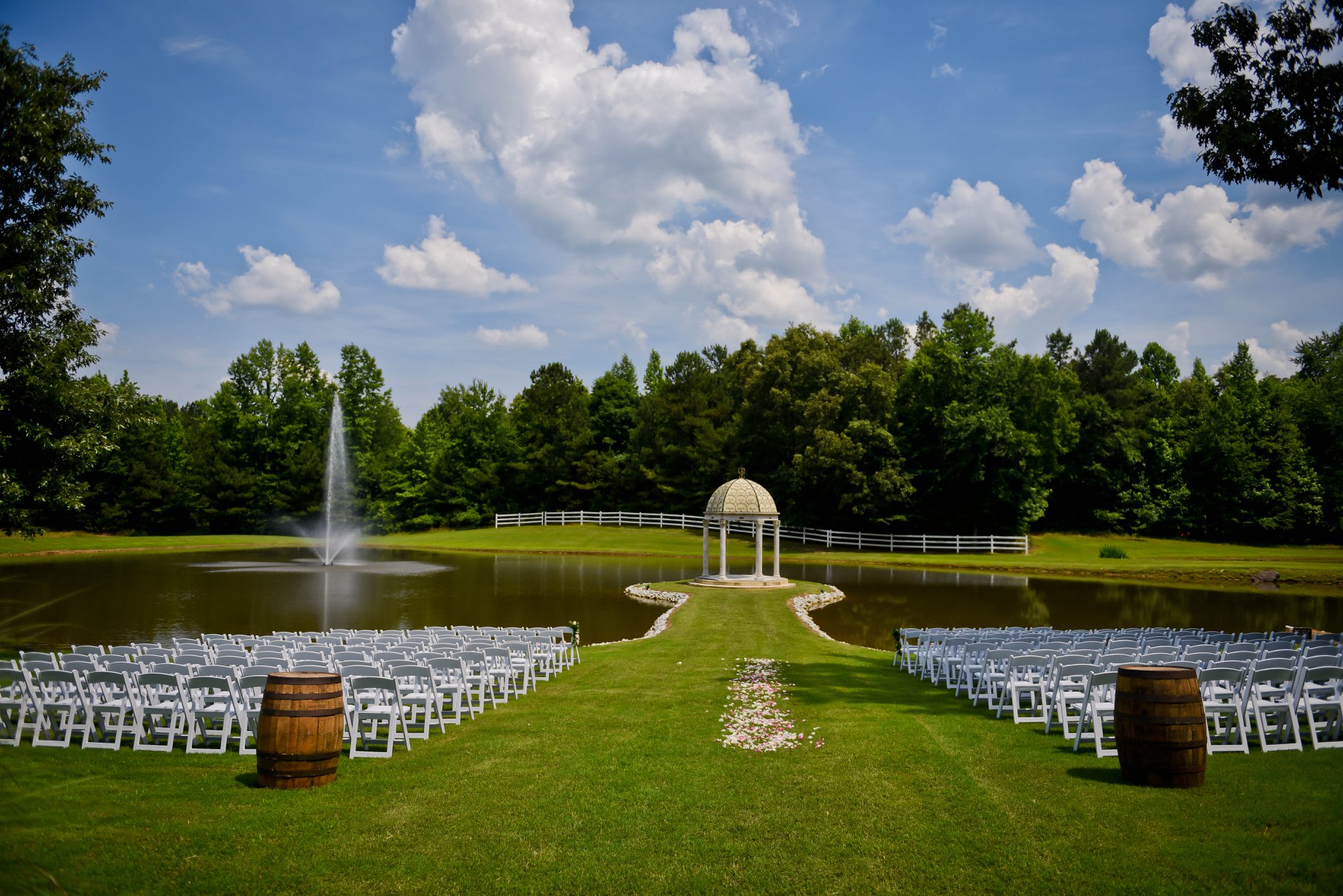 Outdoor wedding venue featuring white chairs arranged facing a gazebo by a pond. A fountain adds elegance to the serene landscape.