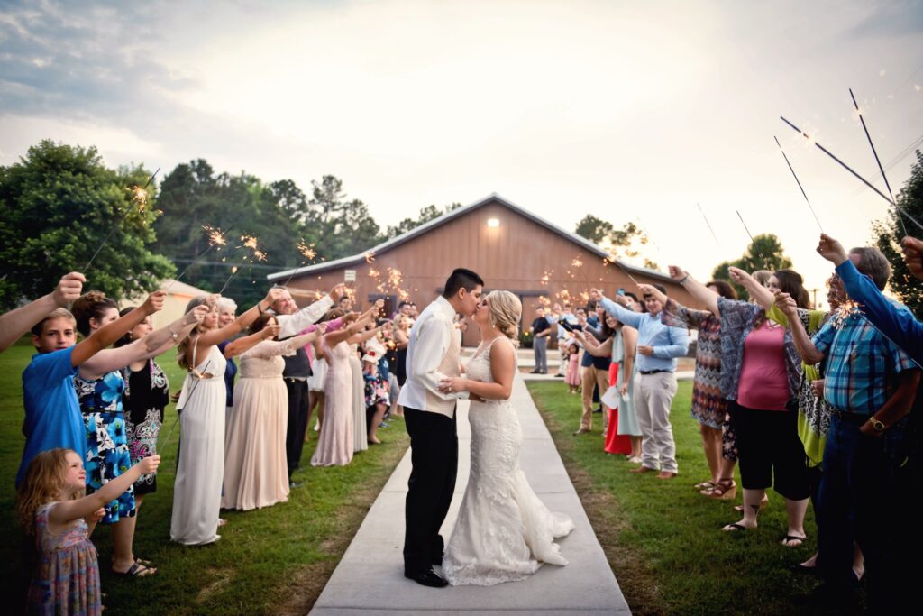 A bride and groom kiss surrounded by wedding guests holding sparklers, celebrating the couple's departure. The scene captures joy and festivity.