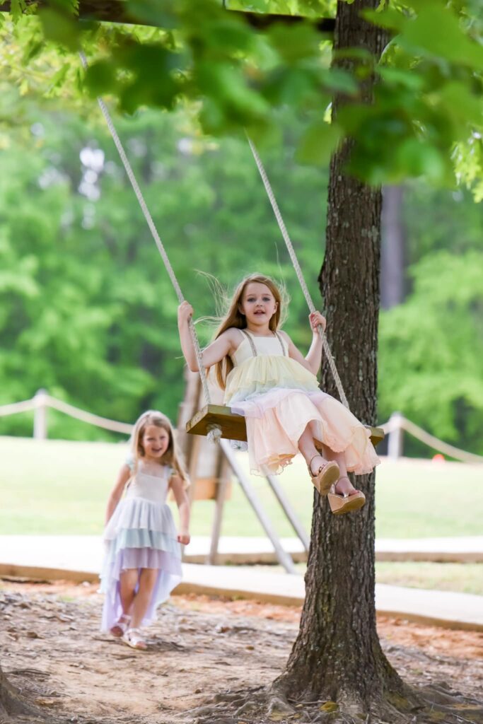 A girl swings joyfully from a tree while a younger girl walks nearby in a park. Lush greenery creates a cheerful, playful atmosphere.
