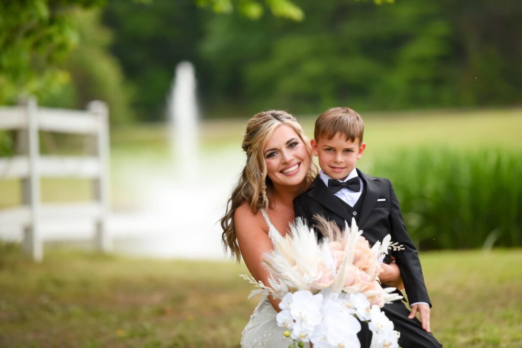 Smiling bride in a white gown hugs a young boy in a tuxedo, both posing with a bouquet by a serene pond and fountain.
