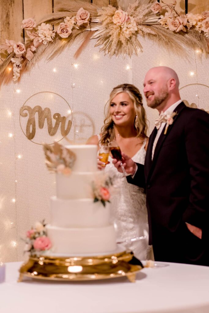 Bride and groom celebrate with a wedding cake adorned with flowers, smiling in front of a decorated backdrop featuring "Mr." in gold.