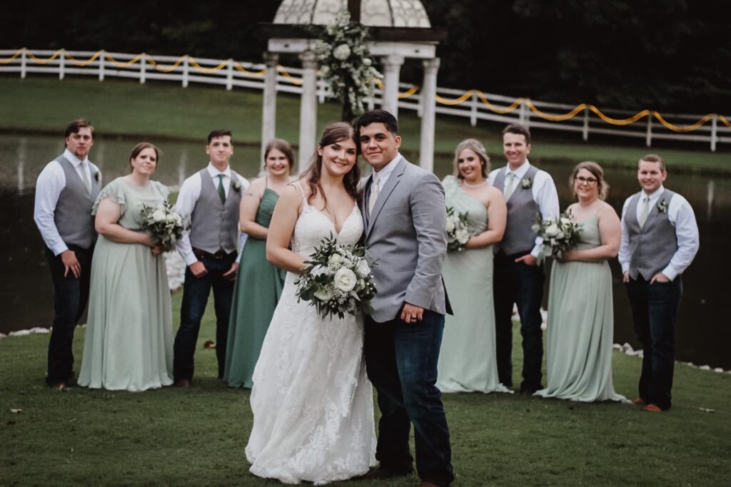 A bride and groom pose in front of their wedding party by a lake, surrounded by greenery and decor, capturing a joyful moment of celebration.