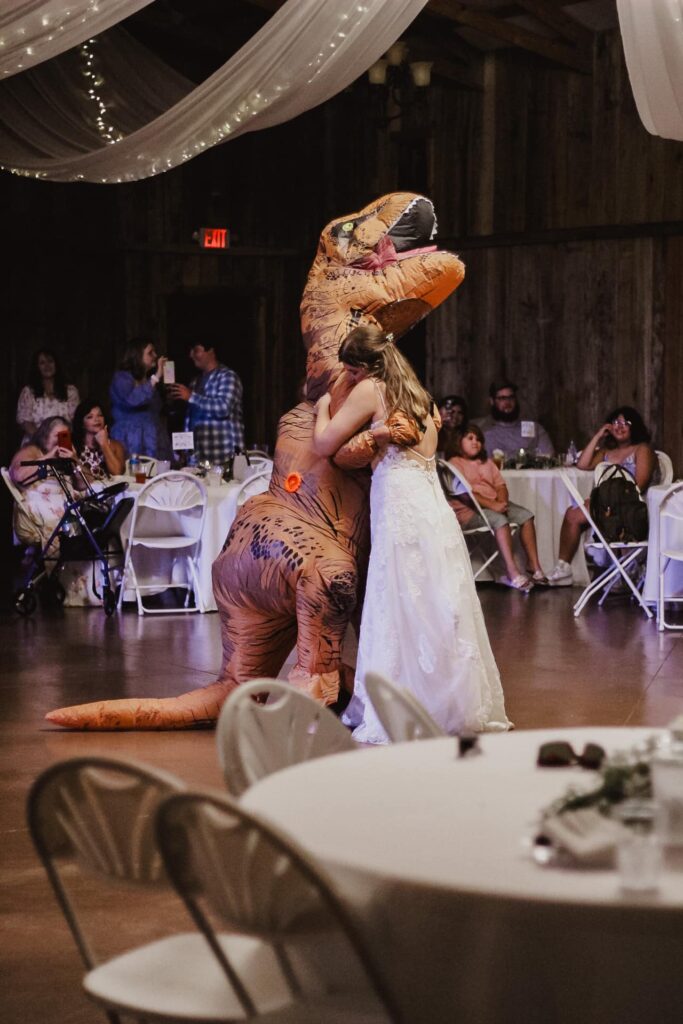 A bride in a white gown hugs a person in a T-Rex inflatable costume at a wedding reception, surrounded by laughing guests and festive decor.