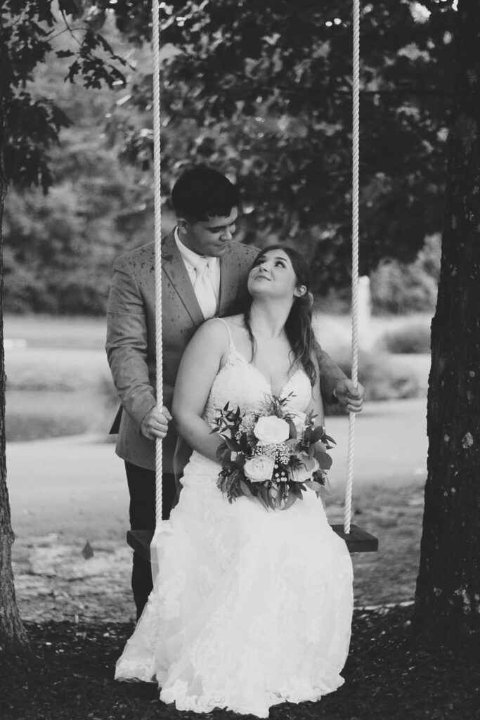 A bride in a white lace gown sits on a swing, gazing up at her groom, who stands behind her, both surrounded by trees.