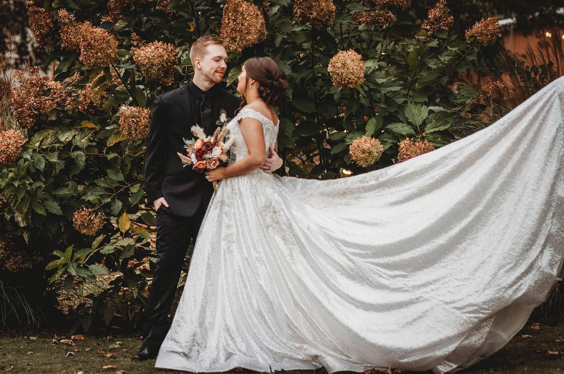 A couple stands embraced in a garden, dressed in formal attire. The bride’s flowing, sparkly gown contrasts with the lush greenery and flowering backdrop.
