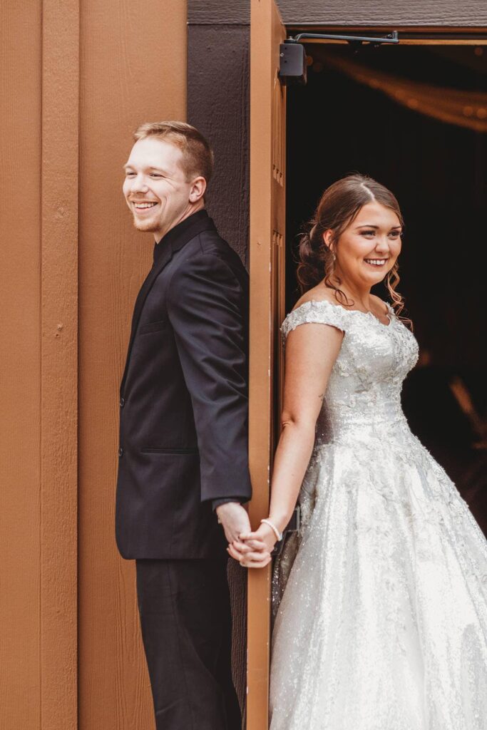 A bride in a sparkling gown and a groom in a black suit hold hands around a doorway, sharing a joyful moment before their wedding.