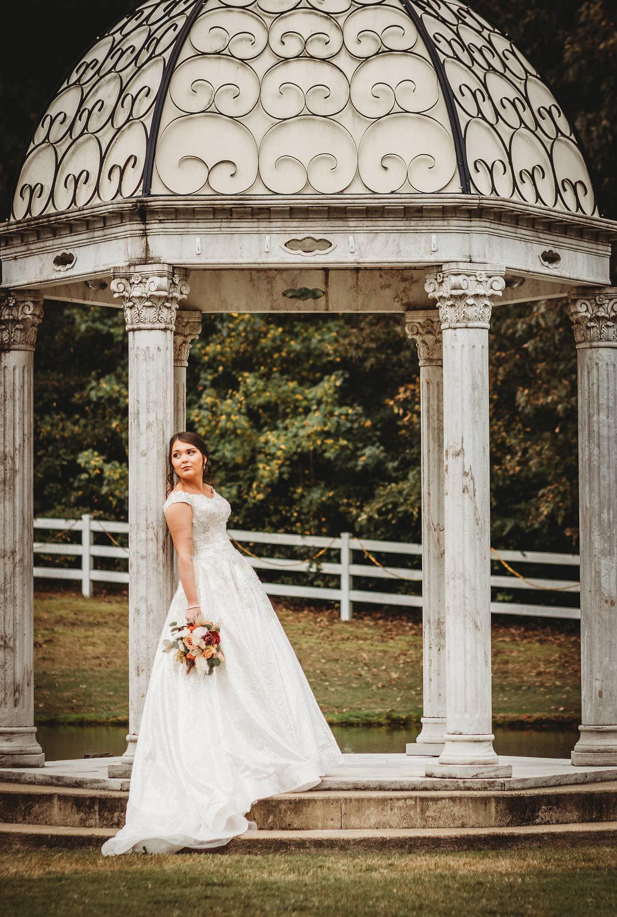 Bride in a elegant white gown stands under a decorative gazebo, holding a bouquet, with lush greenery in the background, capturing a romantic wedding moment.