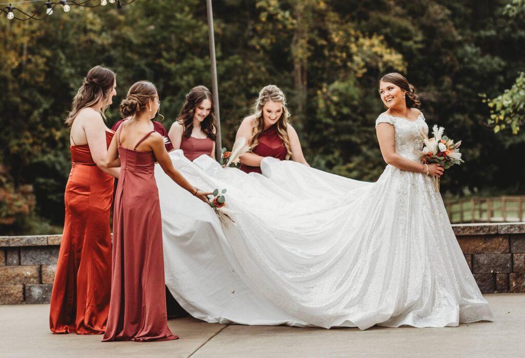 Bridesmaids help the bride with her elegantly flowing white gown in a festive outdoor setting, capturing a moment of joyful preparation before the wedding.