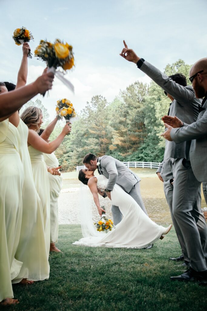 A joyful wedding kiss between the bride and groom, surrounded by cheering guests in pastel dresses and suits, with vibrant flower bouquets.
