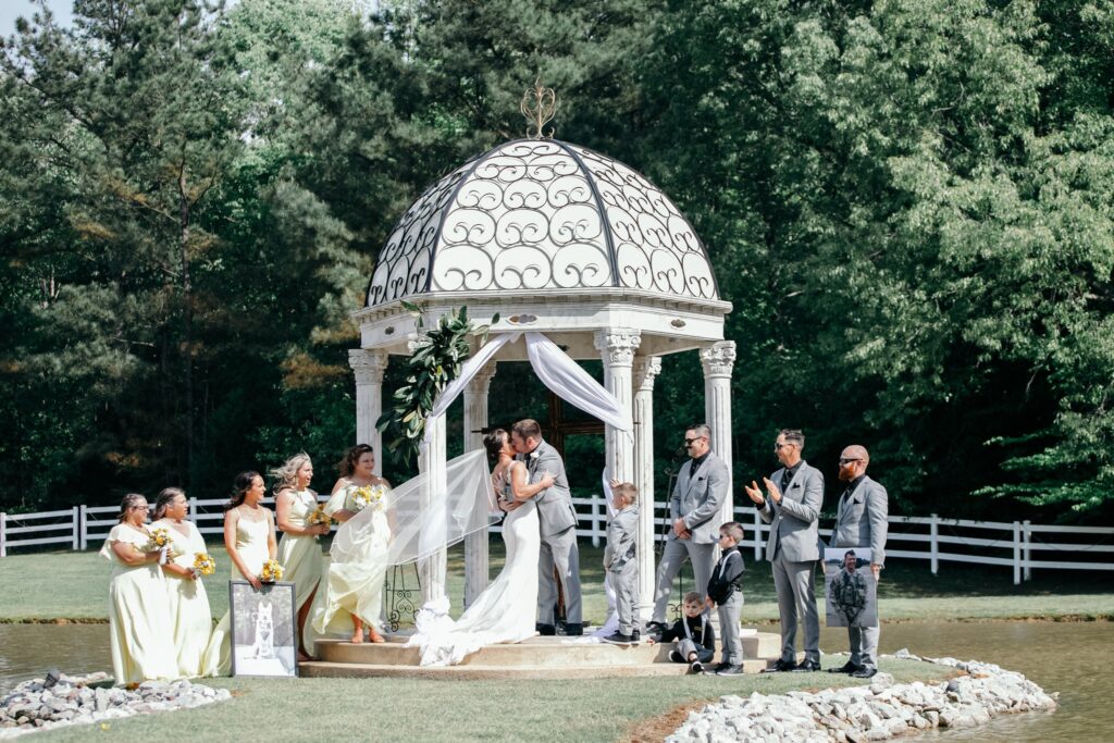 A couple shares a kiss under an ornate gazebo during their outdoor wedding ceremony, surrounded by their bridal party and guests in a lush garden.