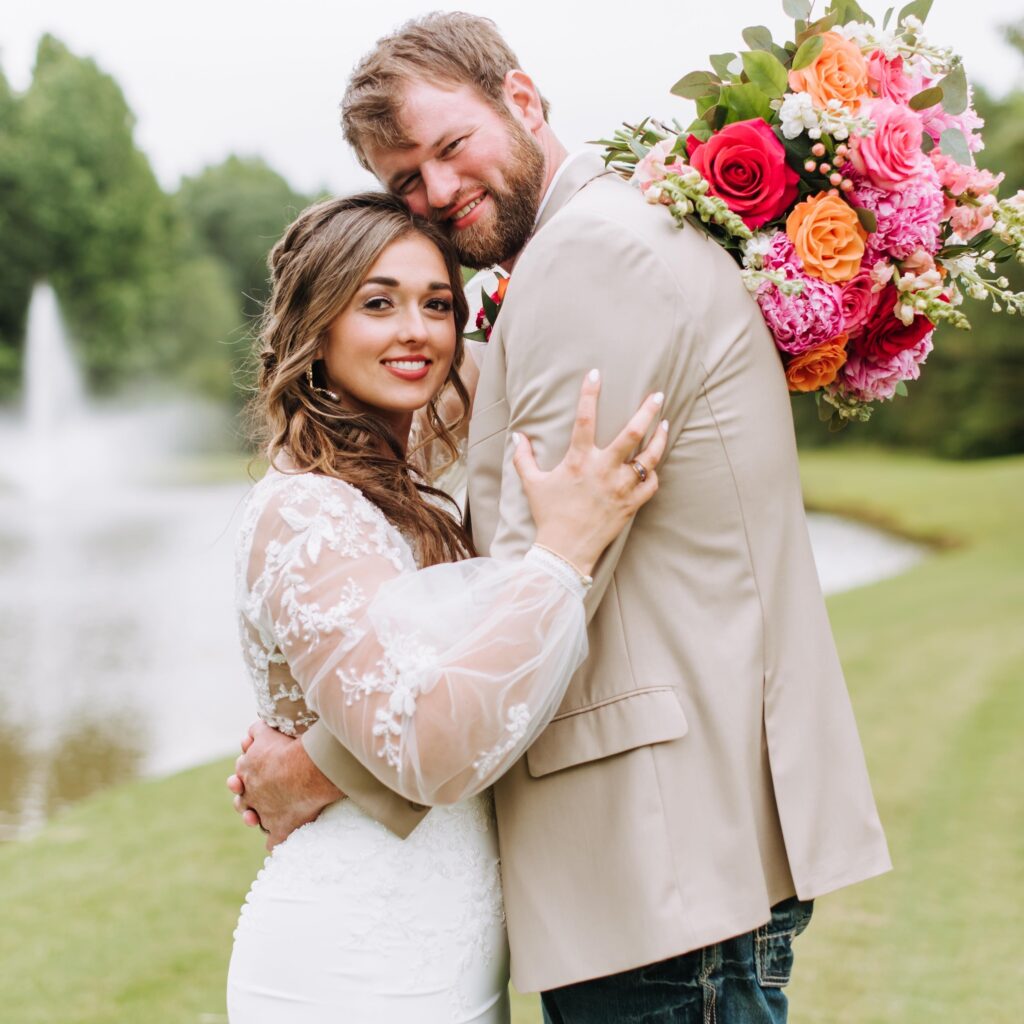 A joyful bride in an embroidered gown embraces her groom, who holds a vibrant bouquet, against a scenic pond backdrop, capturing their wedding bliss.