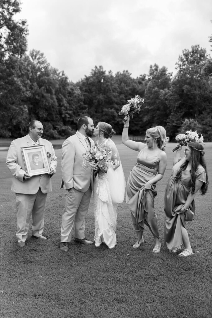 Bride and groom share a kiss while surrounded by their wedding party, holding flowers and a framed portrait, in a vibrant outdoor setting.