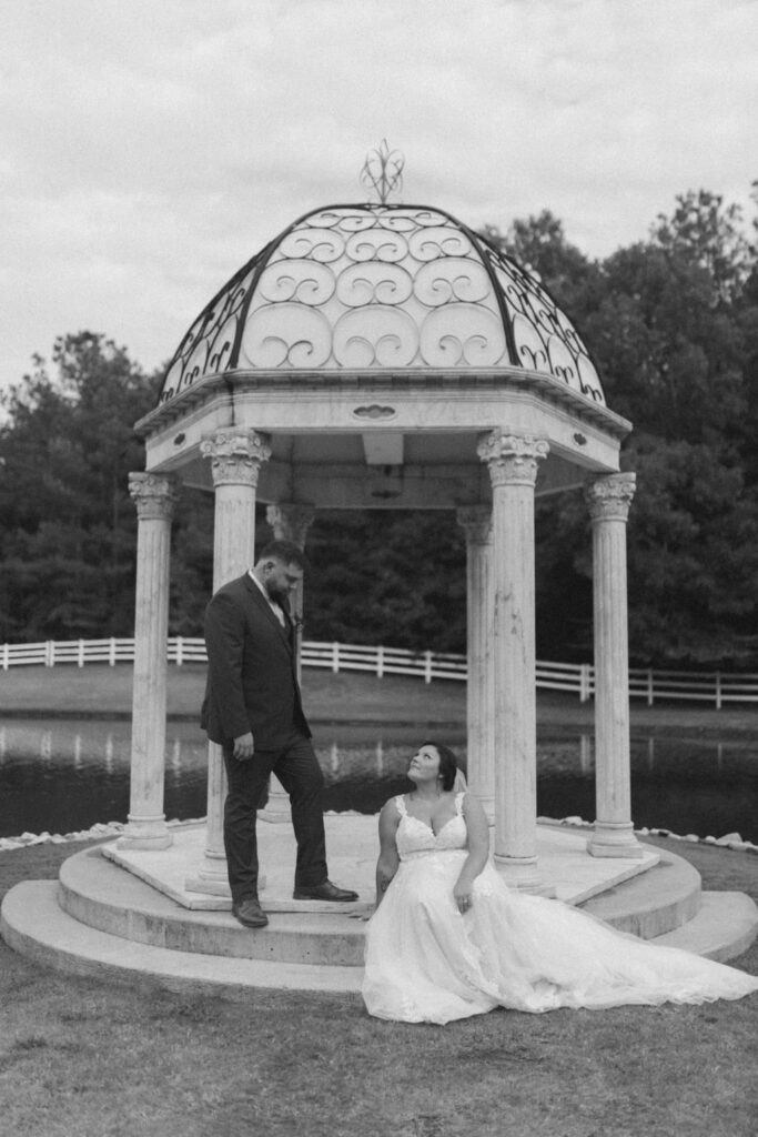 A couple poses romantically in a gazebo near a pond, showcasing their love in an elegant, timeless black-and-white photograph.